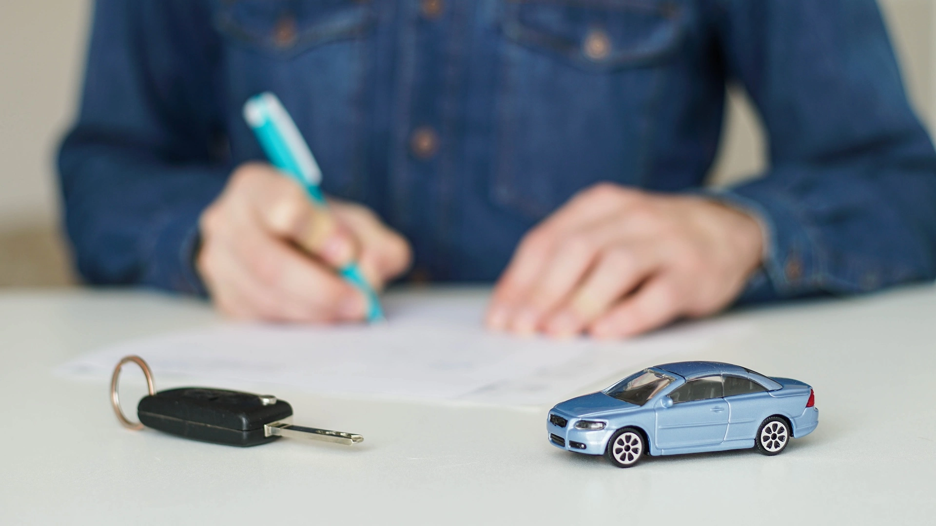 Person completing a car loan application after late payments, with a car key and miniature vehicle.
